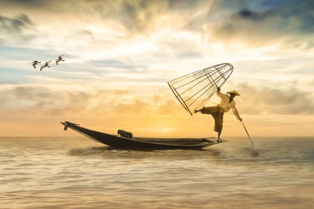 Fisherman balancing on boat with net and oar at sunset, silhouetted against vibrant sky, birds flying nearby.