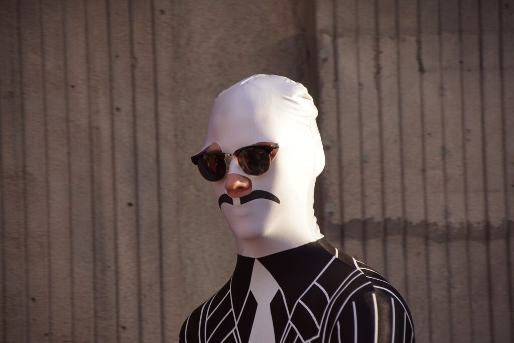 Person in a full-body suit with a mustache design and sunglasses, standing against a textured wall.