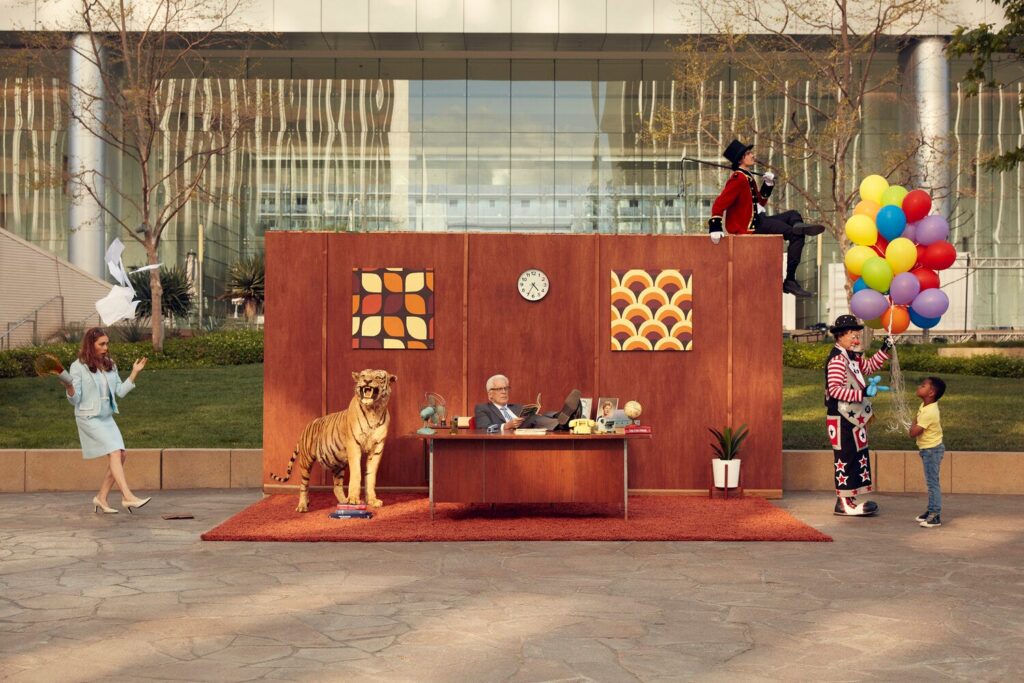 Surreal outdoor office scene with people, a faux tiger, clown, and balloons in front of a modern building.