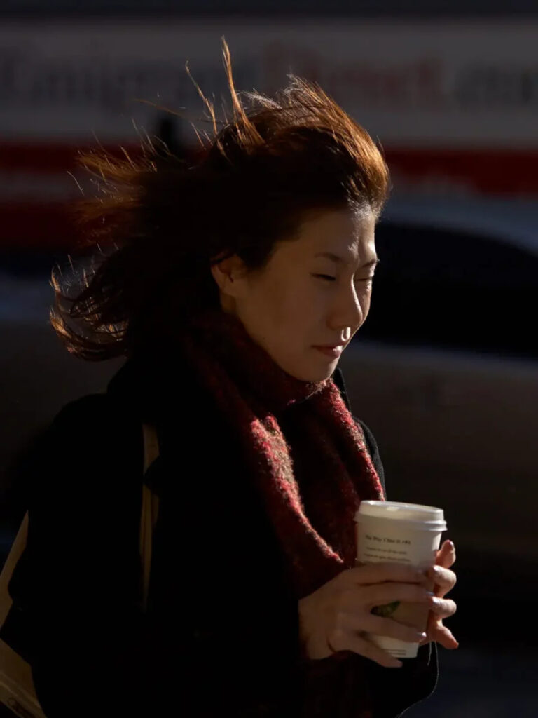 Person holds coffee cup, standing against wind with hair blowing. Sunlit face, wearing a red scarf.