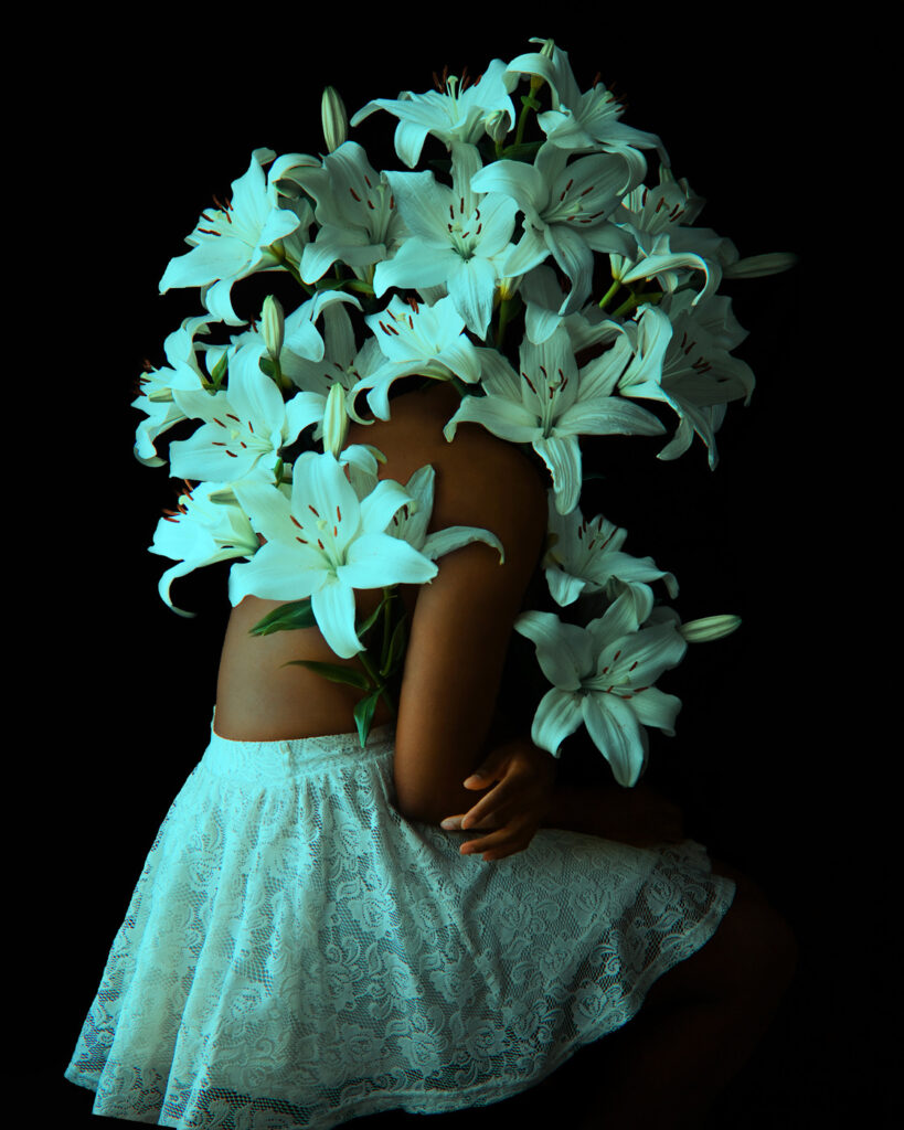 A person with a bouquet of white lilies covering their head, wearing a lace skirt against a black background.