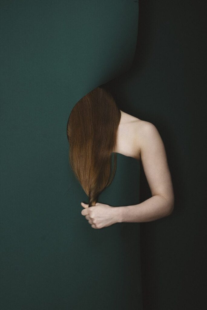 Person with long brown hair peeking through a dark green fabric, pulling their hair with their hand.