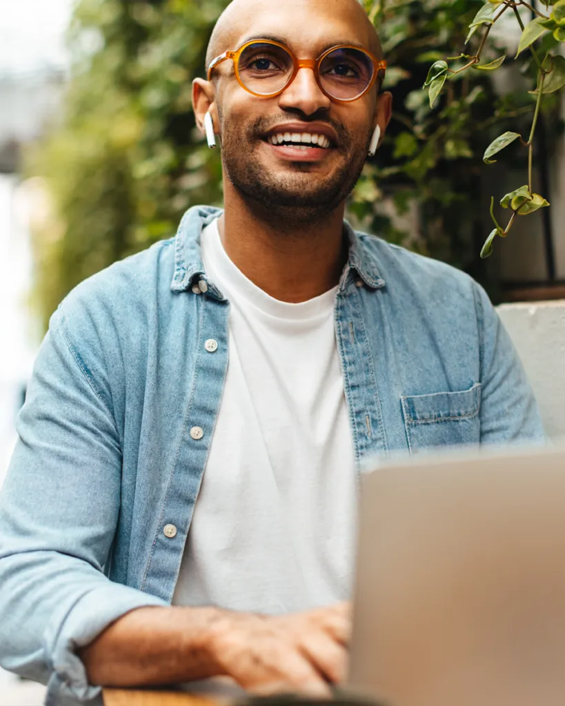 Smiling person with glasses and earbuds, wearing a denim shirt, working on a laptop outdoors near greenery.