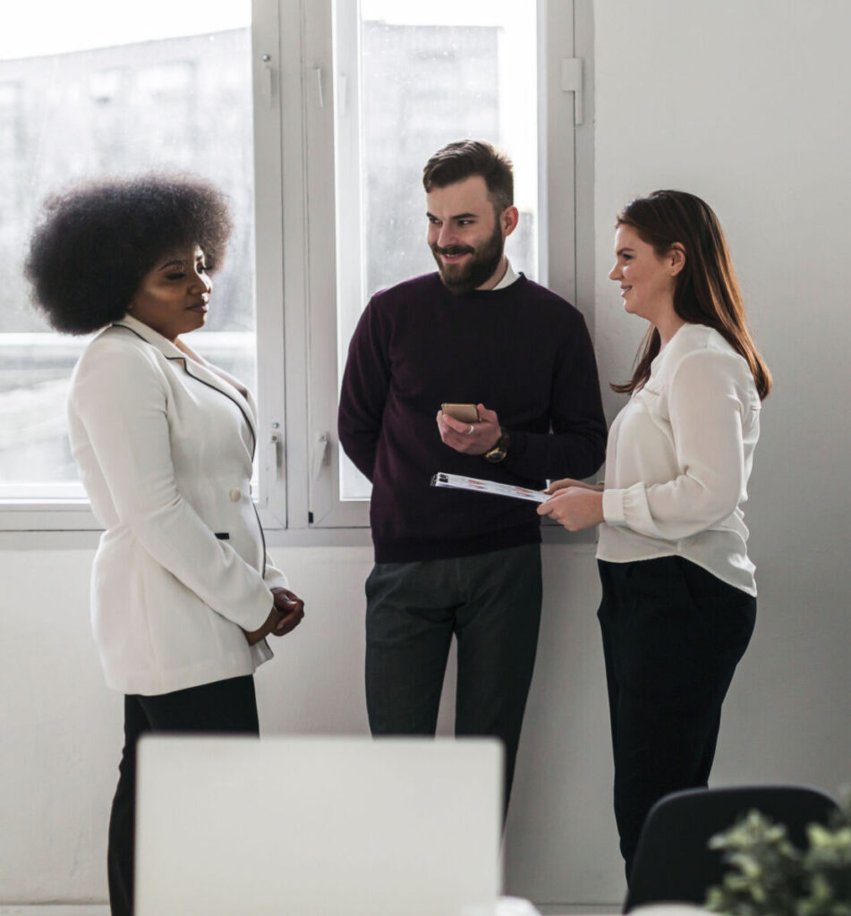 Three colleagues in an office setting, engaged in a conversation near a window, one holding a clipboard.