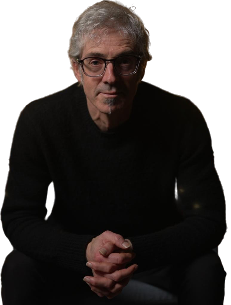 Elderly person with glasses and silver hair, seated in a dark sweater, hands clasped, looking toward the camera.