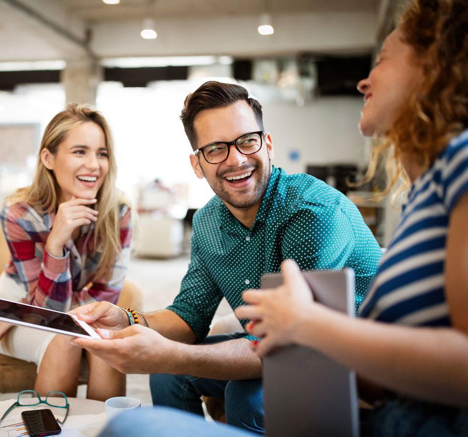A group of three smiling people engaging in conversation, one holding a tablet, in a bright, modern office setting.
