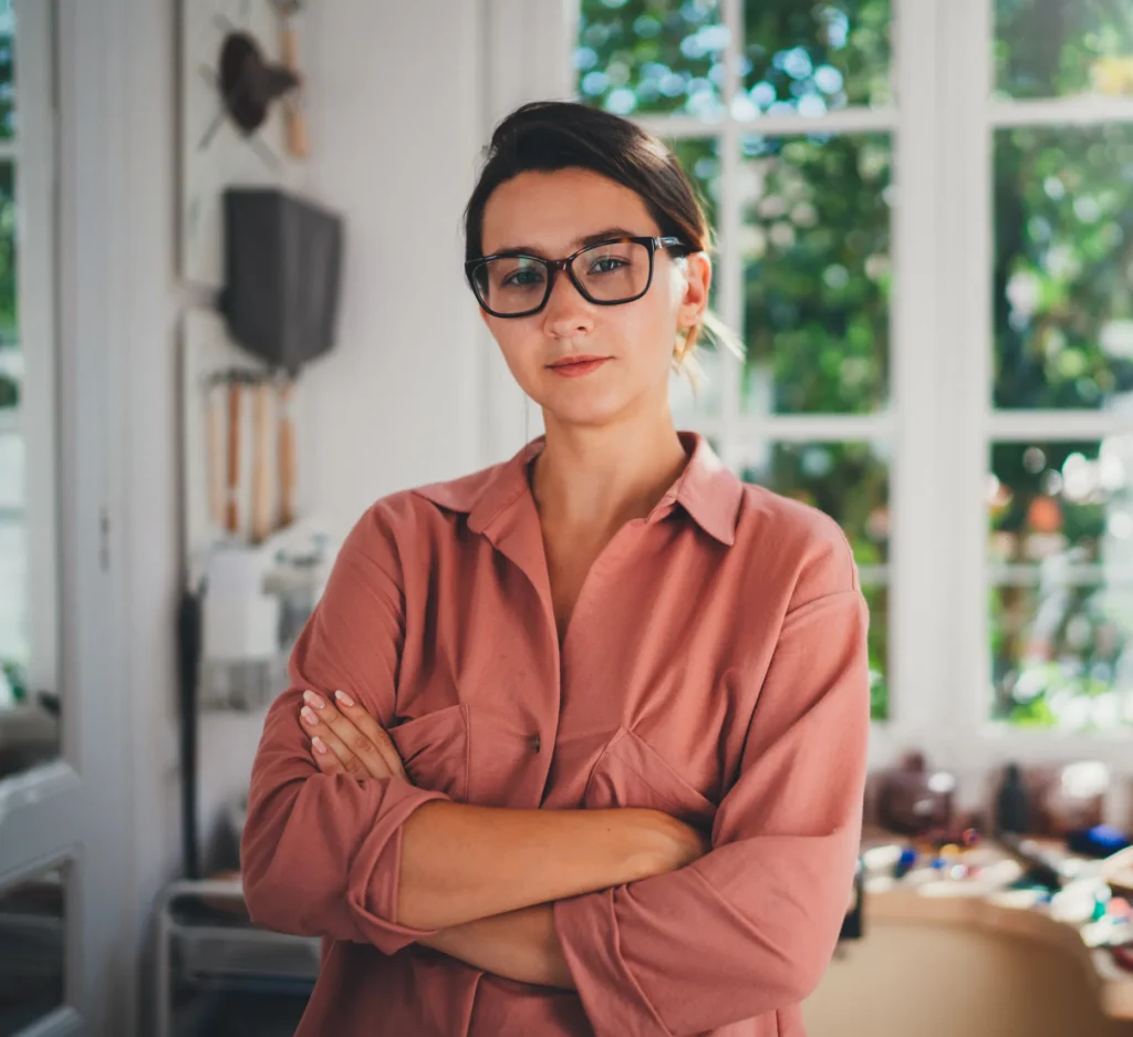 Confident woman in glasses and a pink shirt stands in a bright workspace, arms crossed, with tools and windows behind her.