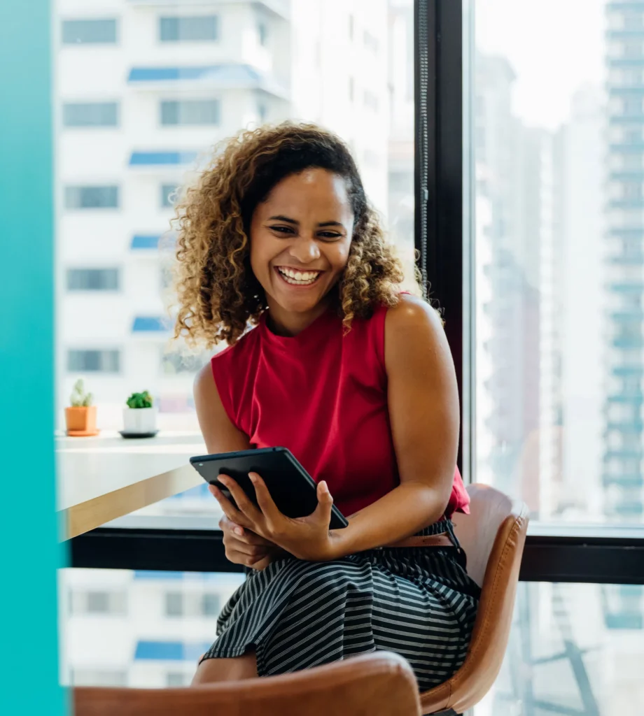 Smiling woman in red top sitting by window with tablet, cityscape background.