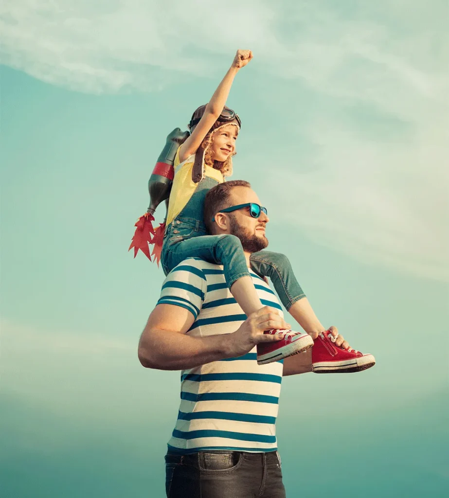 Man in striped shirt carries child with toy jetpack on shoulders, child raises fist triumphantly against a blue sky.