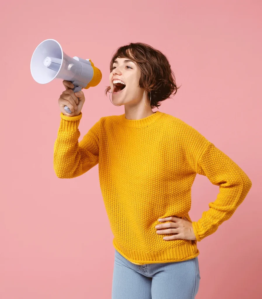 Young woman with short hair, wearing a yellow sweater, shouting into a megaphone against a pink background.