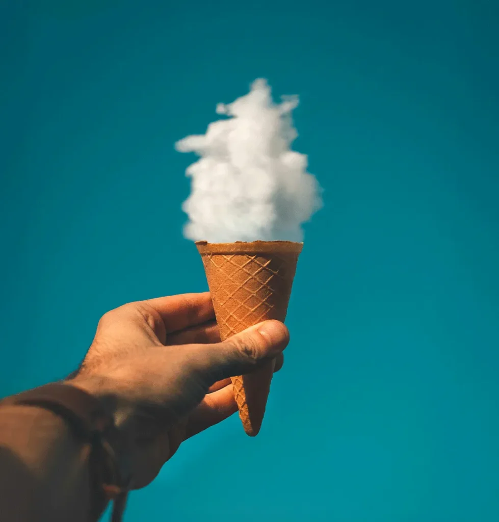 Hand holding an ice cream cone with a cloud-like scoop against a bright blue sky background.