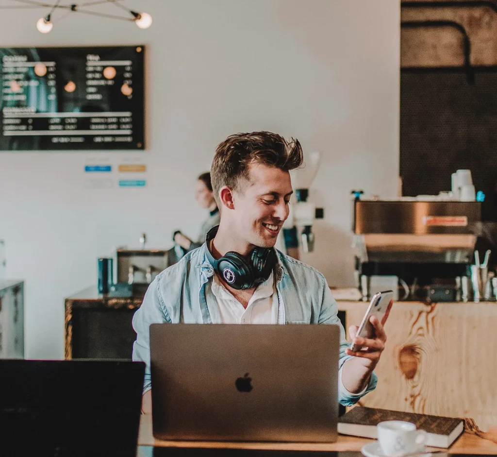 Man smiling at phone, wearing headphones, sitting with laptop in cozy coffee shop.